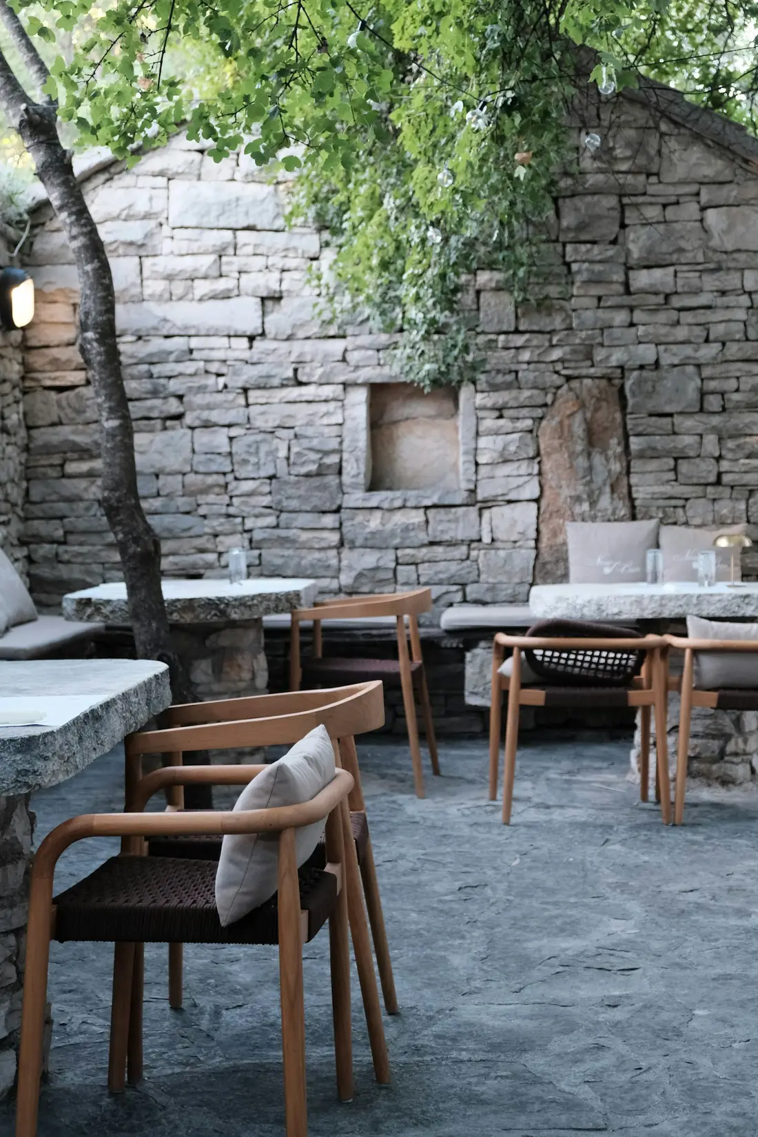Terrasse ombragée contre un mur de pierre, table et chaises en bois.